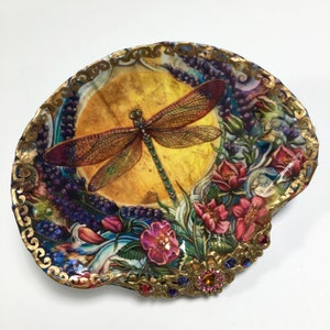 May include: A decorative seashell dish with a colorful dragonfly and floral design. The shell is accented with gold paint and rhinestones.