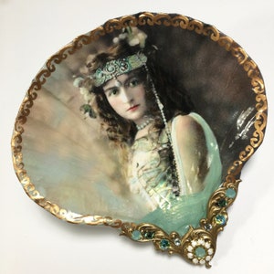 May include: A vintage portrait of a woman with a jeweled headband and pearl necklace is displayed in a decorative seashell with a gold trim and green and white embellishments.