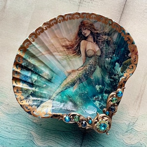 May include: A decorative seashell featuring a mermaid illustration. The artwork depicts a mermaid with flowing auburn hair and a scaled tail, set against an underwater scene. The shell is edged with gold trim and adorned with blue and green gemstones.