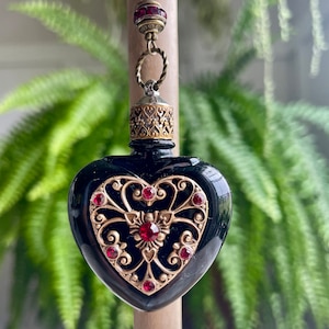 May include: A black heart-shaped glass bottle with a gold filigree design and red rhinestone accents hangs from a gold chain with a red rhinestone bead.
