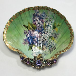 May include: A green seashell dish with a gold trim and a floral design of purple and white lilacs. The dish has a decorative gold and jeweled trim.