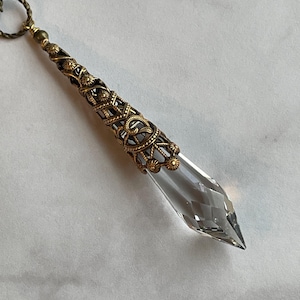 May include: A clear crystal point pendant with a gold-toned filigree design. The pendant is attached to a brown cord.