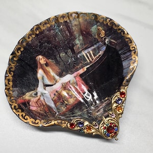 May include: A decorative seashell dish with a gold trim and a painted image of a woman in a boat. The dish is decorated with colorful rhinestones.