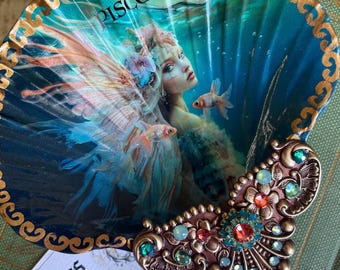 Può includere: Una conchiglia decorativa con un designo di sirena e pesci. La conchiglia è dipinta in tonalità di blu, verde e oro. Il testo "Pisces" è scritto sulla conchiglia. La conchiglia è decorata con un ornamento in oro e rame.