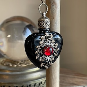 May include: A black heart-shaped glass bottle with a gold filigree design and a red gemstone. The bottle is hanging from a gold chain with a silver bead.