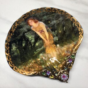 May include: A decorative seashell dish with a gold trim and a painted image of a woman in a forest setting. The dish has a jeweled embellishment on the bottom edge.