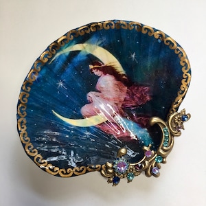 May include: A blue seashell with a gold trim and a decorative image of a woman sitting on a crescent moon. The seashell has a gold metal handle with blue and purple rhinestones.