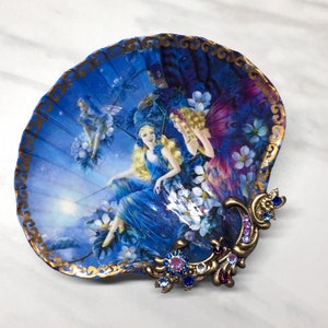 Puede incluir: Un plato de concha azul y dorado con un diseño floral y tres hadas. El plato tiene un borde dorado y un adorno de joyas.