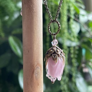 May include: A delicate necklace featuring a pendant shaped like a pink flower bud. The pendant is encased in a bronze-colored metal with intricate leaf details. The chain is also bronze-colored, and the background is blurred green foliage.