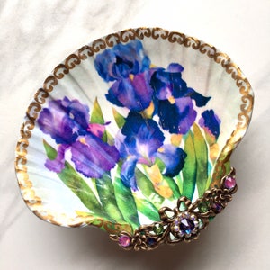 May include: A white seashell dish with a gold trim and a floral design of purple irises. The dish has a decorative gold and jeweled trim.