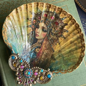 May include: A decorative seashell featuring a painted portrait of a woman wearing an ornate headdress. The shell has a gold and teal color scheme with a jeweled metal accent. The artwork has a vintage, bohemian aesthetic.