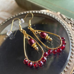 May include: Gold-toned teardrop earrings with deep red faceted beads. The earrings feature a delicate wire design, with a cluster of red beads at the bottom and a single bead at the top. The earrings are displayed on a reflective surface.