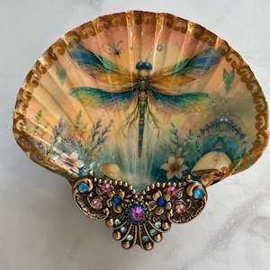 May include: A seashell with a painted dragonfly design and gold accents. The shell is decorated with a jeweled trim.