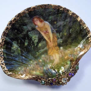 May include: A seashell dish with a gold trim and a painted image of a woman in a forest with glowing lights. The dish is decorated with green, purple, and clear rhinestones.