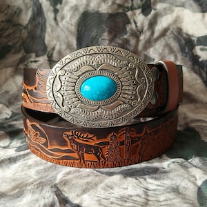 以下が含まれることがあります： ターコイズ色の石が付いた装飾的なシルバーバックルが付いた茶色の革ベルト。ベルトには、ワシ、鹿、木を含む自然のシーンがエンボス加工されています。バックルは楕円形です。