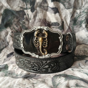 以下が含まれることがあります： 黒い革のベルトに、銀と金のサソリの装飾バックルが付いています。ベルトにはエンボス加工された花柄があります。バックルは黒い背景に銀色の縁取りがあり、中央に金のサソリがいます。