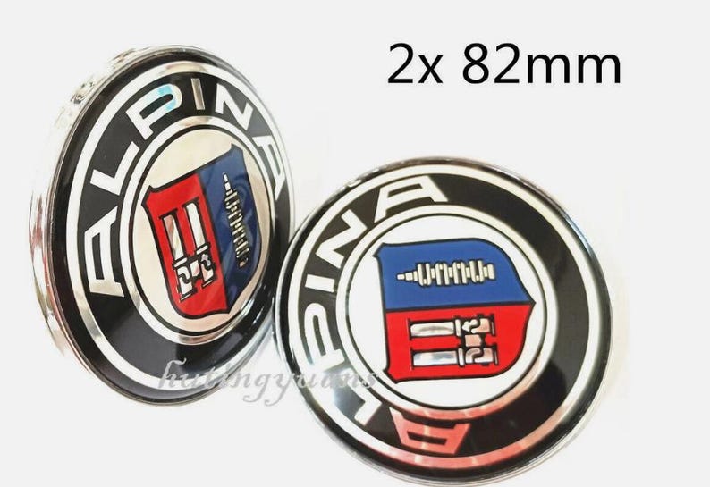 K&ouml;nnte beinhalten: Zwei Alpina-Radnabenabdeckungen. Jede Kappe ist rund, schwarz und silberfarben und tr&auml;gt das Alpina-Logo in der Mitte. Das Logo zeigt ein blau-rotes Schilddesign. Der Text "2x 82mm" ist sichtbar.