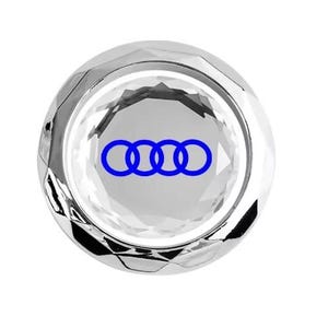 May include: A clear, faceted crystal car emblem featuring the Audi logo in blue. The emblem is circular and has a reflective, polished surface. The Audi logo consists of four interlocking rings.