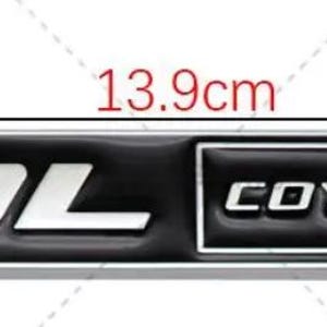 May include: A black and silver rectangular emblem with the text "5.0L COYOTE V8". The emblem is 13.9cm long and 2cm high, with a silver border. The text is in a bold, sans-serif font.