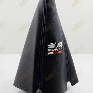Peut inclure: Soufflet de levier de vitesses en cuir noir avec surpiqûres bleues. Le soufflet a une surface texturée et une garniture argentée en haut. Le mot "POWER" est brodé en blanc et orange, avec des caractères japonais au-dessus.