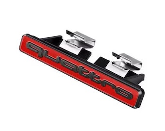 May include: A red and black Quattro emblem with silver mounting clips. The emblem features the word "Quattro" in black lettering on a red background. The mounting clips are silver.