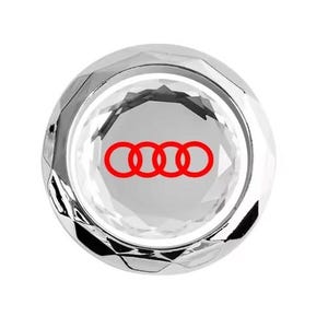 Op de afbeelding: Een cirkelvormig auto-embleem van helder, facetgeslepen kristal, met het Audi-logo in rood. Het embleem heeft een gepolijst, reflecterend oppervlak. Het Audi-logo bestaat uit vier in elkaar grijpende ringen.