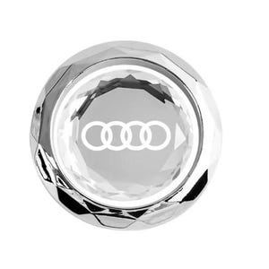 May include: A clear, faceted crystal car emblem with the Audi logo in white. The emblem is circular and has a reflective, metallic appearance. The Audi logo consists of four interlocking rings.