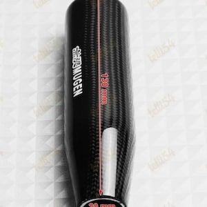 Può includere: Un pomello del cambio in fibra di carbonio nera, lungo 130 mm, largo 33 mm e con un diametro di 20 mm. Il pomello ha un motivo intrecciato e la scritta "MUGEN" in bianco.