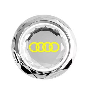 May include: A faceted, silver-colored emblem featuring the Audi logo, a set of four interlocking yellow rings. The emblem has a reflective, jewel-like appearance. The background is white.