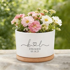 May include: A white ceramic planter with a wooden base, filled with pink and white flowers. The planter has the word "Engaged" and the date "05.18.25" in black script, with a heart symbol.