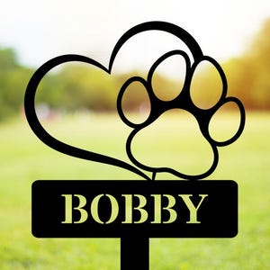 May include: A black metal garden stake featuring a heart outline and a paw print silhouette above a rectangular nameplate with the name BOBBY cut out in capital letters. The design is mounted on a vertical post for placement in the ground.
