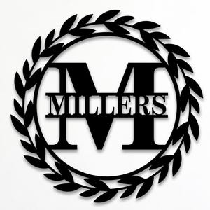 Puede incluir: Un letrero de pared circular negro con un borde de corona de hojas de laurel que rodea una letra M mayúscula grande con el nombre MILLERS centrado horizontalmente sobre la letra. El texto Custom Order es visible en la esquina superior izquierda.