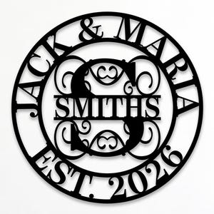 Puede incluir: Un letrero de pared circular de metal negro con los nombres JACK & MARIA, el apellido SMITHS y el texto EST. 2026. El diseño incluye una letra S central grande con patrones de volutas decorativas dentro de un marco circular.