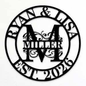 Könnte beinhalten: Ein kreisförmiges Wandobjekt aus schwarzem Metall mit den Namen RYAN & LISA im oberen Bogen, dem Nachnamen MILLER in der Mitte, dem Buchstaben M und dem Text EST. 2026 am unteren Rand. Das Design enthält dekorative Schnörkel-Elemente.