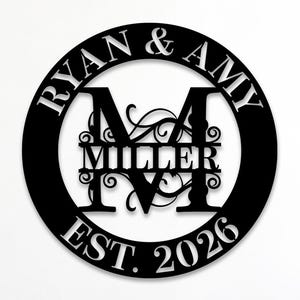 Puede incluir: Un letrero de pared circular de metal negro que presenta los nombres RYAN & AMY en la parte superior, una letra M central grande, el nombre MILLER en el medio y el texto EST. 2026 en la parte inferior, con adornos decorativos de volutas.