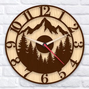 Könnte beinhalten: Eine runde Wanduhr aus Holz mit Berg- und Waldmotiv. Das Zifferblatt hat einen hellen Holzmaserungshintergrund mit dunkelbraunen Ausschnitten. Die Uhrzeiger sind weiß mit einem roten Sekundenzeiger.