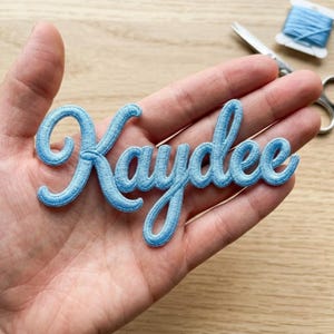 May include: A light blue embroidered patch featuring the name Kaydee in a cursive script font. The patch is made of dense thread stitching and is designed to be applied to fabric surfaces for personalisation or decorative purposes.