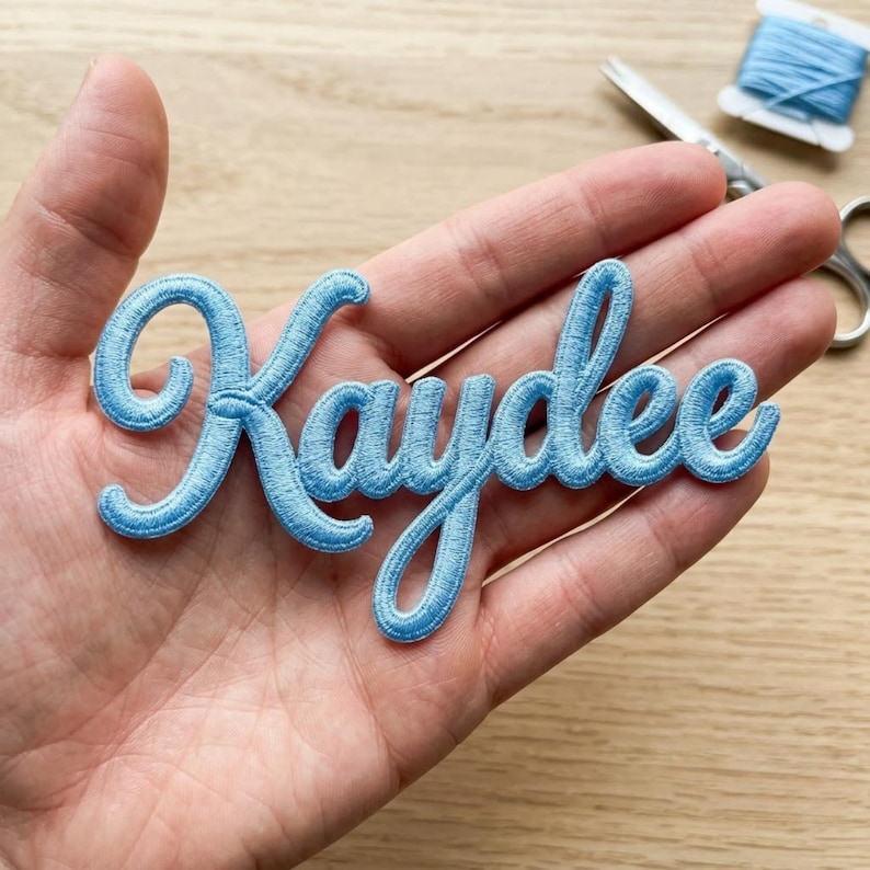 May include: A light blue embroidered patch featuring the name Kaydee in a cursive script font. The patch is made of dense thread stitching and is designed to be applied to fabric surfaces for personalisation or decorative purposes.
