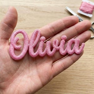 May include: A light pink embroidered patch featuring the name Olivia in a cursive script font. The lettering is stitched with a raised, textured thread finish, creating a distinct outline for each letter in the name.