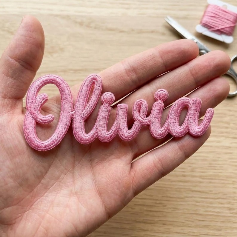 May include: A light pink embroidered patch featuring the name Olivia in a cursive script font. The lettering is stitched with a raised, textured thread finish, creating a distinct outline for each letter in the name.
