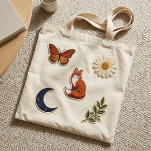 May include: A cream-coloured canvas tote bag featuring five embroidered patches: an orange monarch butterfly, a white daisy, a red fox, a dark blue crescent moon with small gold stars, and a green leafy branch.