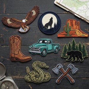 May include: A collection of embroidered patches featuring a bald eagle, a howling wolf in a circular frame, a desert landscape with a cactus, a brown cowboy boot, a teal pickup truck, a black bear in a pine forest, a rattlesnake, and two crossed axes.