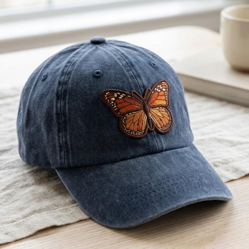 May include: A dark blue baseball cap featuring an embroidered monarch butterfly patch on the front. The butterfly has orange and brown wings with white accents. The cap has a structured crown, a curved brim, and stitched eyelets.