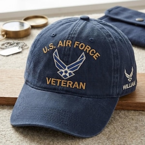 May include: A navy blue baseball cap featuring the embroidered text U.S. AIR FORCE VETERAN in gold thread above a blue and white Air Force emblem. The side of the cap displays a smaller Air Force emblem above the embroidered name WILLIAM in white.