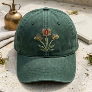 May include: A forest green baseball cap featuring an embroidered design of three Venus flytrap plants. The plants have light green stems and leaves with reddish-brown interiors and small, spiky teeth along the edges of the open traps.
