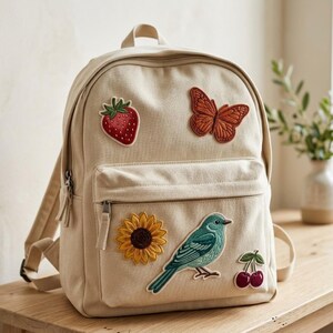 May include: A beige canvas backpack featuring embroidered patches of a red strawberry, an orange butterfly, a yellow sunflower, a blue bird, and two red cherries. The backpack has a main compartment and a front zippered pocket with metal pulls.