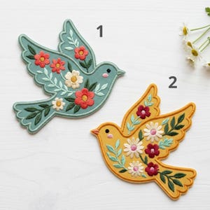 May include: Two embroidered bird patches labeled 1 and 2. Patch 1 is sage green with red and white flowers. Patch 2 is mustard yellow with dark red and white flowers. Both feature green leaf embroidery and are shaped like birds in flight.