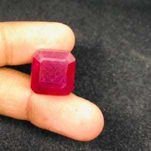 Op de afbeelding: Een vierkant geslepen robijn edelsteen met facetten. De edelsteen heeft een diepe, verzadigde rode kleur, met meerdere facetten die het licht vangen. De robijn wordt tussen de vingers gehouden, wat de grootte en vorm laat zien. De achtergrond is een donker, getextureerd oppervlak.