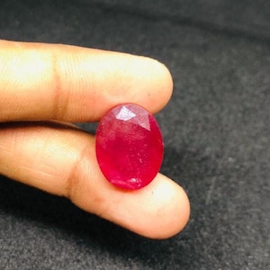 Puede incluir: Una gema de rubí facetada de forma ovalada. La gema es de un color rojo intenso y vibrante, con una superficie pulida que refleja la luz. El rubí se sostiene entre los dedos, mostrando su tamaño y forma.