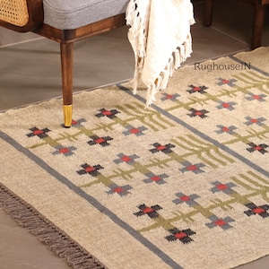 May include: Beige area rug with a geometric pattern featuring red, black, and gray cross-like shapes and green vertical lines. The rug has a gray border and fringe along the edge. The rug is placed near a wooden chair.